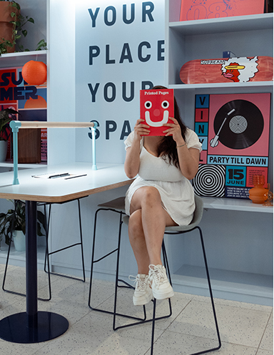 A woman sits at a small table in a brightly lit room, holding a book in front of her face.